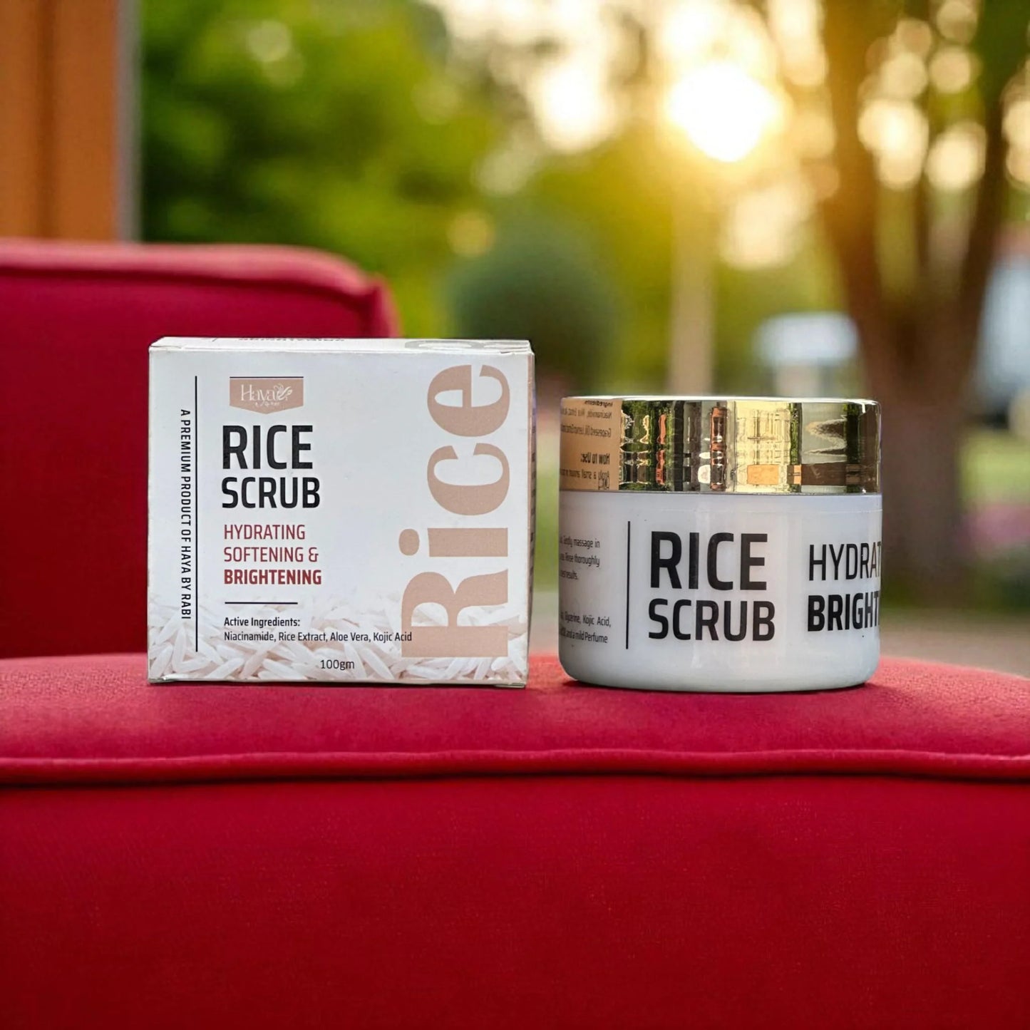 Haya by Rabi Rice Scrub jar and box displayed on a red surface with a soft outdoor background.